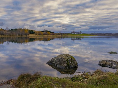 Elliðavatn í logni, skýin speglast í vatninu og í forgrunni eru stórir steinar í vatninu. Elliðabærinn í bakgrunni hinum megin við vatnið.