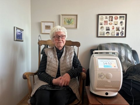 Eldri kona situr í stól og við hlið hennar er lyfjaskammtaratæki