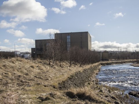 Toppstöðin in Elliðaárdalur, with yellow markings and the Elliðaá River in the foreground and a blue sky with scattered clouds in the background.