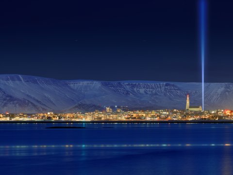 Friðarsúlan. Mynd tekin í myrkri frá borginni, Esjan í baksýn, sjór í forgrunni. Bláir tónar, súlan lengst til hægri.