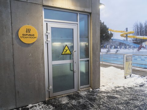 Sauna í Breiðholtslaug, myndin sýnir hana að utan, hurðina inn og skiltið við hlið hurðar. Sundlaugin í bakgrunni. Vetur, snjór.