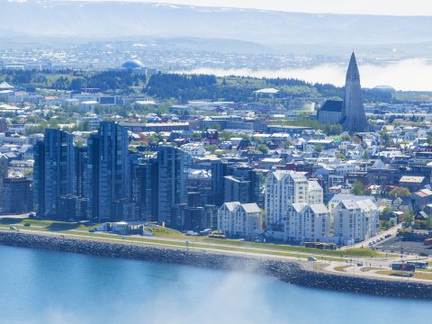 Loftmynd yfir Reykjavík, útsýni frá Sæbraut, yfir miðborg, að Hallrímskirkju og Perlu. Sjór í forgrunni, fjöll og sjór í bakgrunni. Mistur yfir.