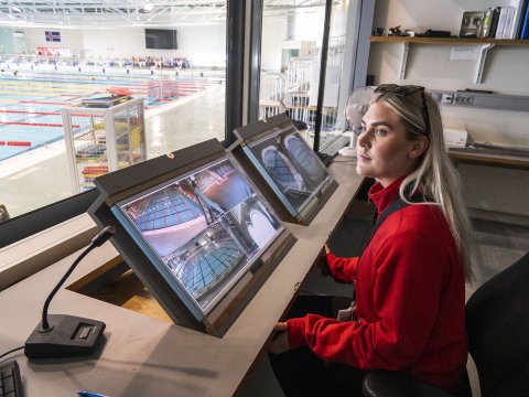 Sundlaugarvörðurinn Dagbjört Ýr Gísladóttir vaktar skjái í vaktherbergi Laugardalslaugar. Situr í rauðri peysu, gluggi út að innilaug vinstra megin í mynd.