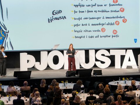 Arna Ýr Sævarsdóttir, Head of Office for services and transformation at the Department of Service & Innovation, at Reykjavík City's management day at Harpa 2024. Arna center stage, with the letters ÞJÓNUSTA prominent on the stage and slides in the background. Seated guests in the foreground.