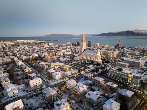 Aerial view over the city center, with Hallgrímskirkja prominent in the center, and Faxaflói Bay and the mountains in the distance. Clear skies, winter light.