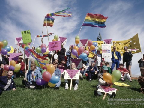 A colorful group of people holding rainbow flags and signs gathers on a grassy area, likely Arnarhóll. Clear weather with a bright blue sky overhead.