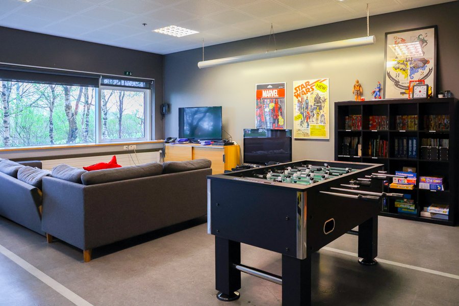 A foosball table, a gray sofa in front of a television. A black shelf and posters on the wall.