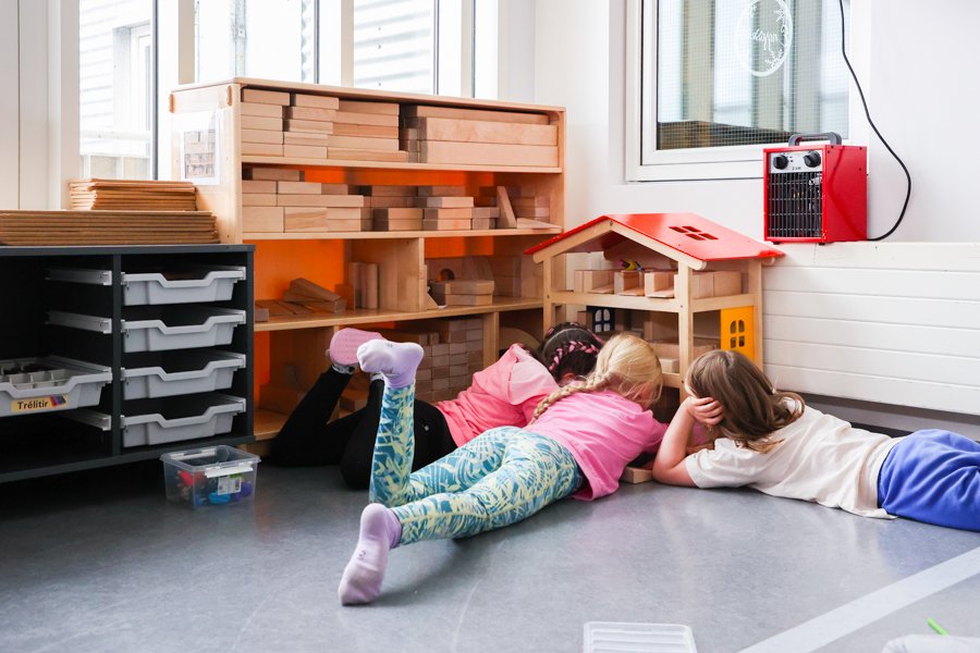 Three children lie on the floor in front of a shelf with large blocks and a small dollhouse. 