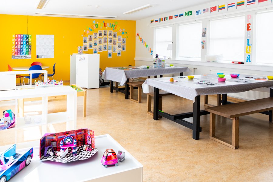 Tables and chairs in a hall. A yellow wall with various decorations and a row of flags above the windows. 
