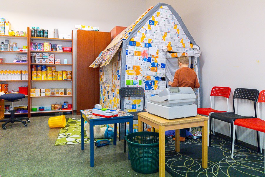 A child entering a small house built from milk cartons. A table, chairs and a cash register in front.