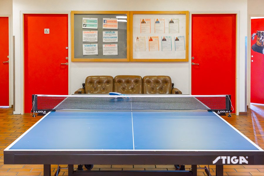 Room with a ping pong table, couch and notices on the wall. Red doors in the background. 