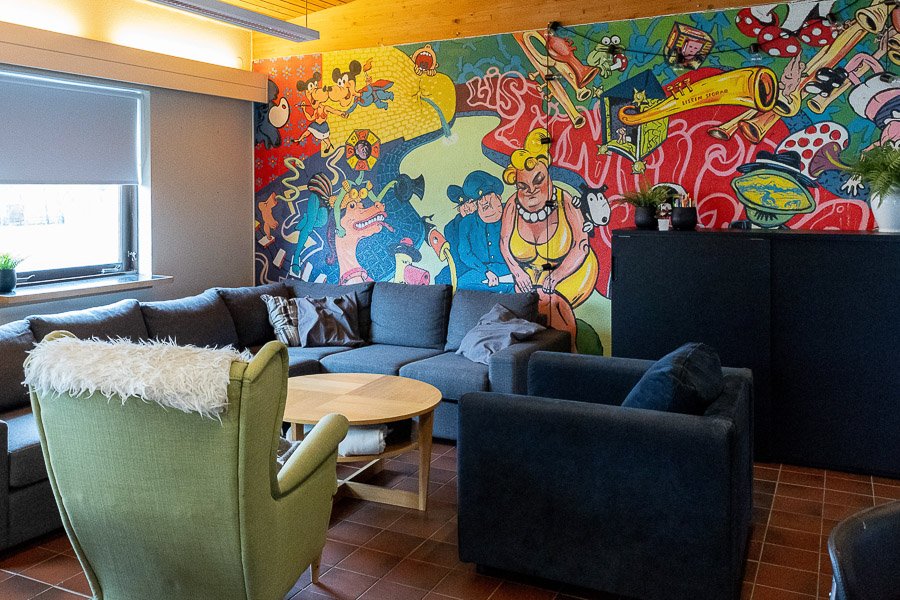 A large mural in the background. A corner couch, a coffee table, and chairs. A black shelf. 