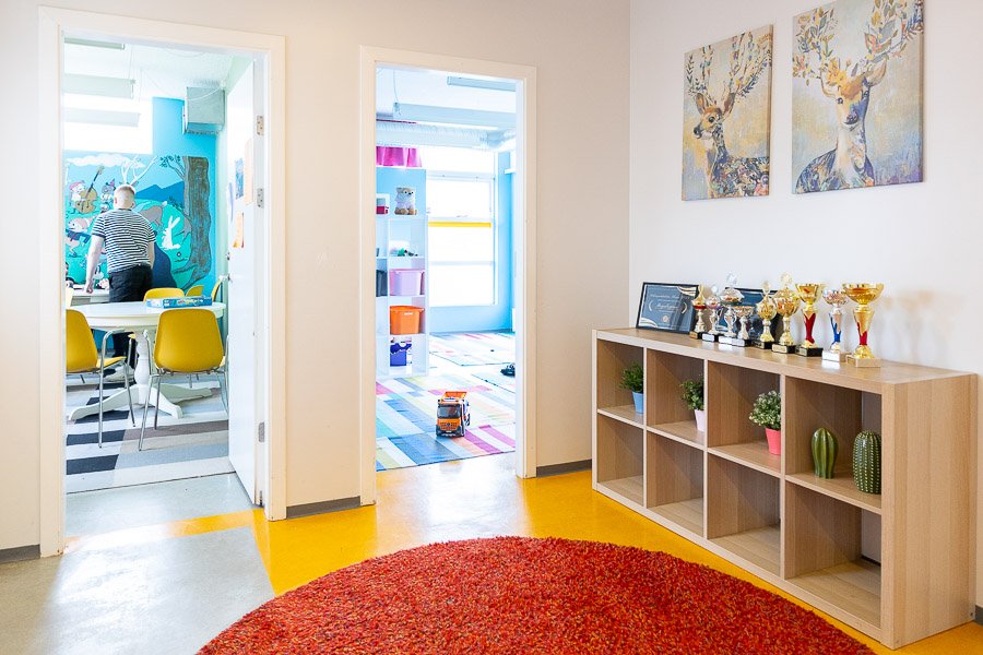 Looking into two rooms from a hallway. The hallway has a red rug and a shelf with awards. In one of the rooms, you can see the back of a child.