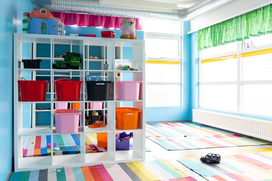 A room with large windows and green curtains on one side. Colorful rugs on the floor and a large shelf in the center of the room with plastic bins in different colors. 
