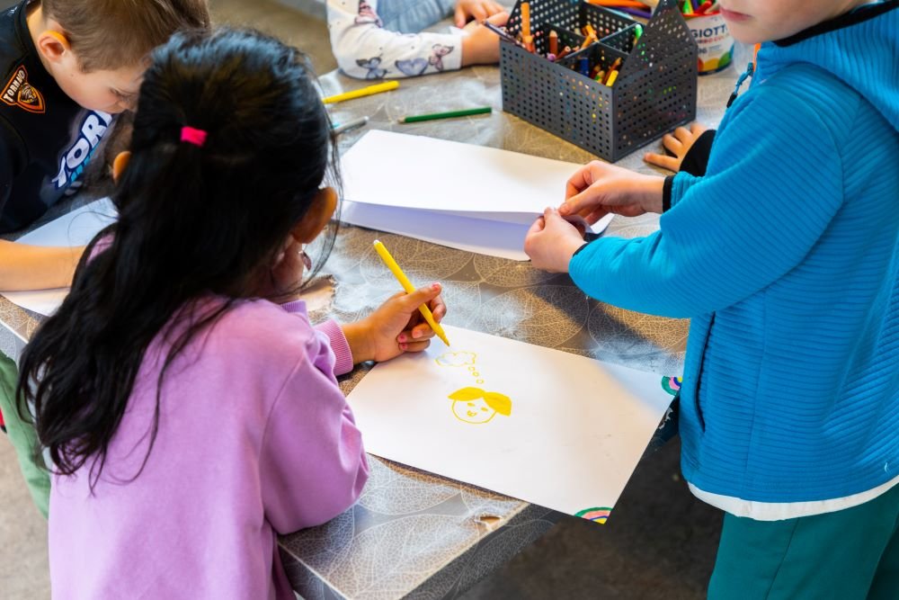 Children sit and stand at a table coloring. 