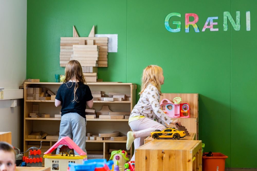 A green wall with the word “Græni” (Green) written on it. A shelf with large wooden blocks and other toys. Children at play. 