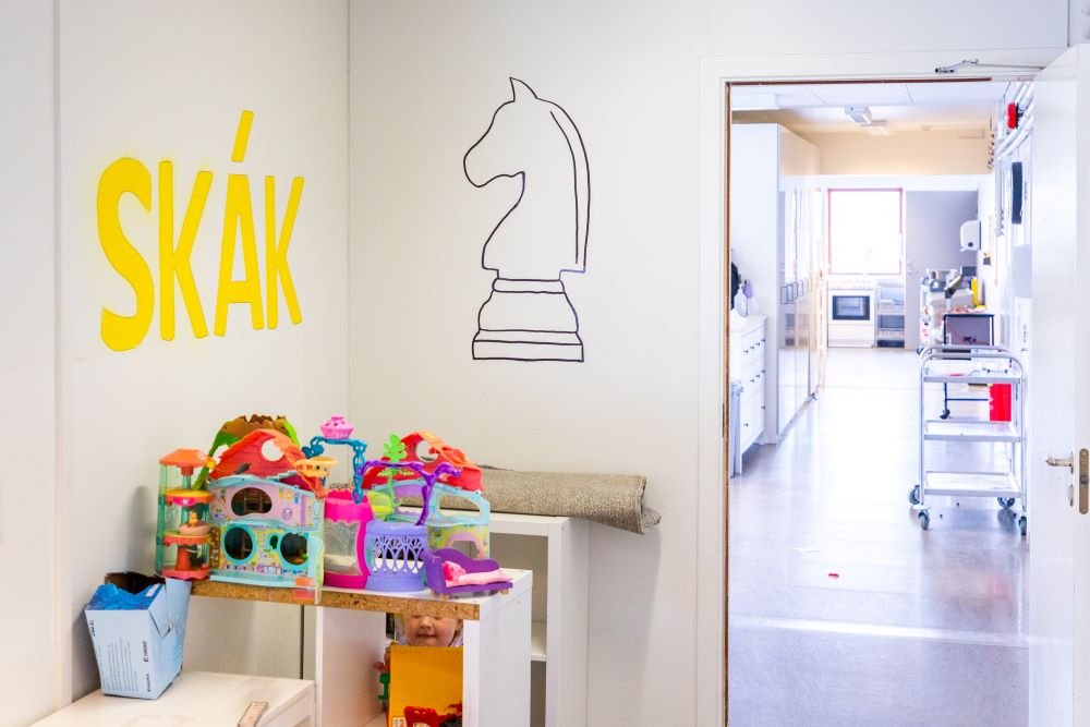 A white-painted room with a picture of a chess piece and the word “Skák” (Chess) on the wall. Toys on a shelf. 