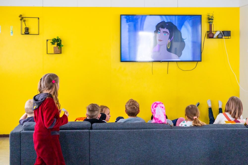 Children sit on a gray sofa watching a cartoon on a screen hanging on a yellow wall.
