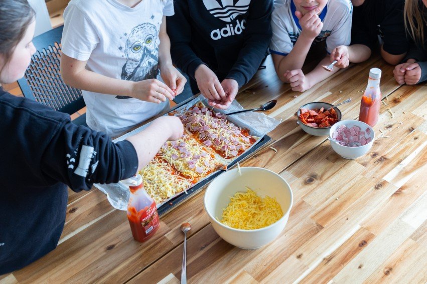 Children stand at a table putting toppings on a pizza.