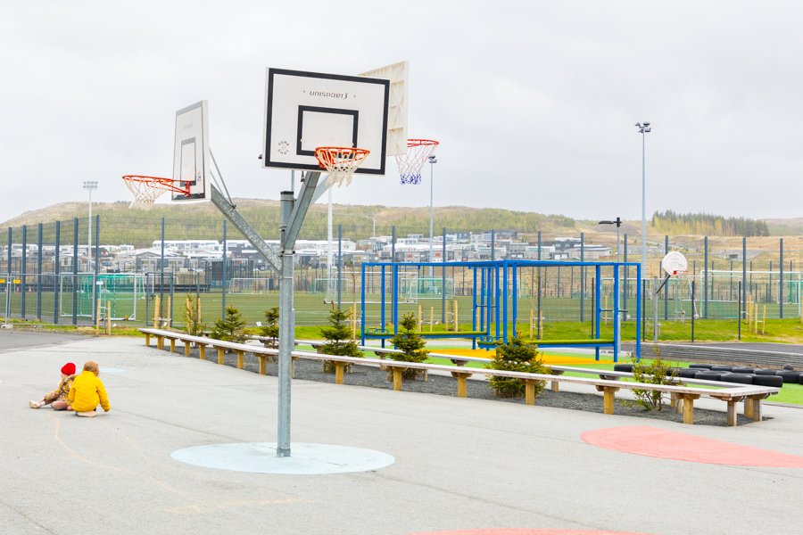 A basketball court with children sitting on the ground. A fenced soccer field in the background.