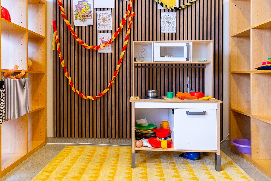 A toy kitchen on a yellow rug. A brown wall with concertina-folded paper decorations in the background. Shelves on both sides. 