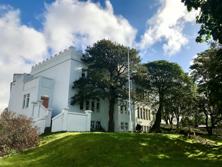 Hús Söngskólans í Reykjavík séð utanfrá. Stór tré í garðinum, hvítt hús og blár himinn.