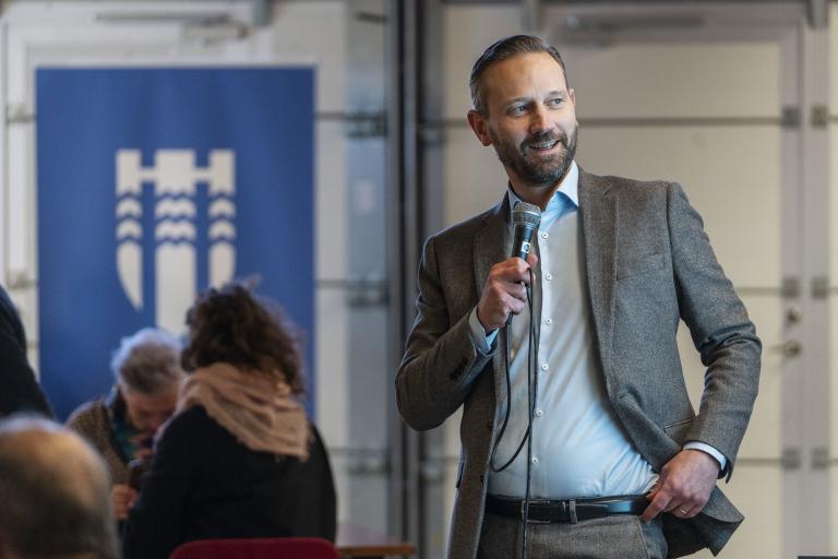 Mayor Einar Þorsteinsson speaks into a microphone at a resident forum. The city's emblem is visible in the background.