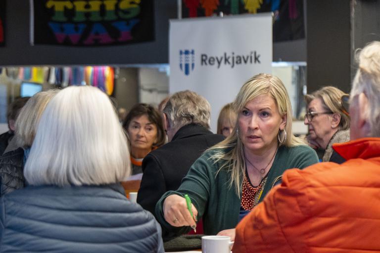 Attendees gather for the Miðborg resident forum at Spennistöðin. A woman stands in the foreground, with the city's emblem visible on a banner in the background.
